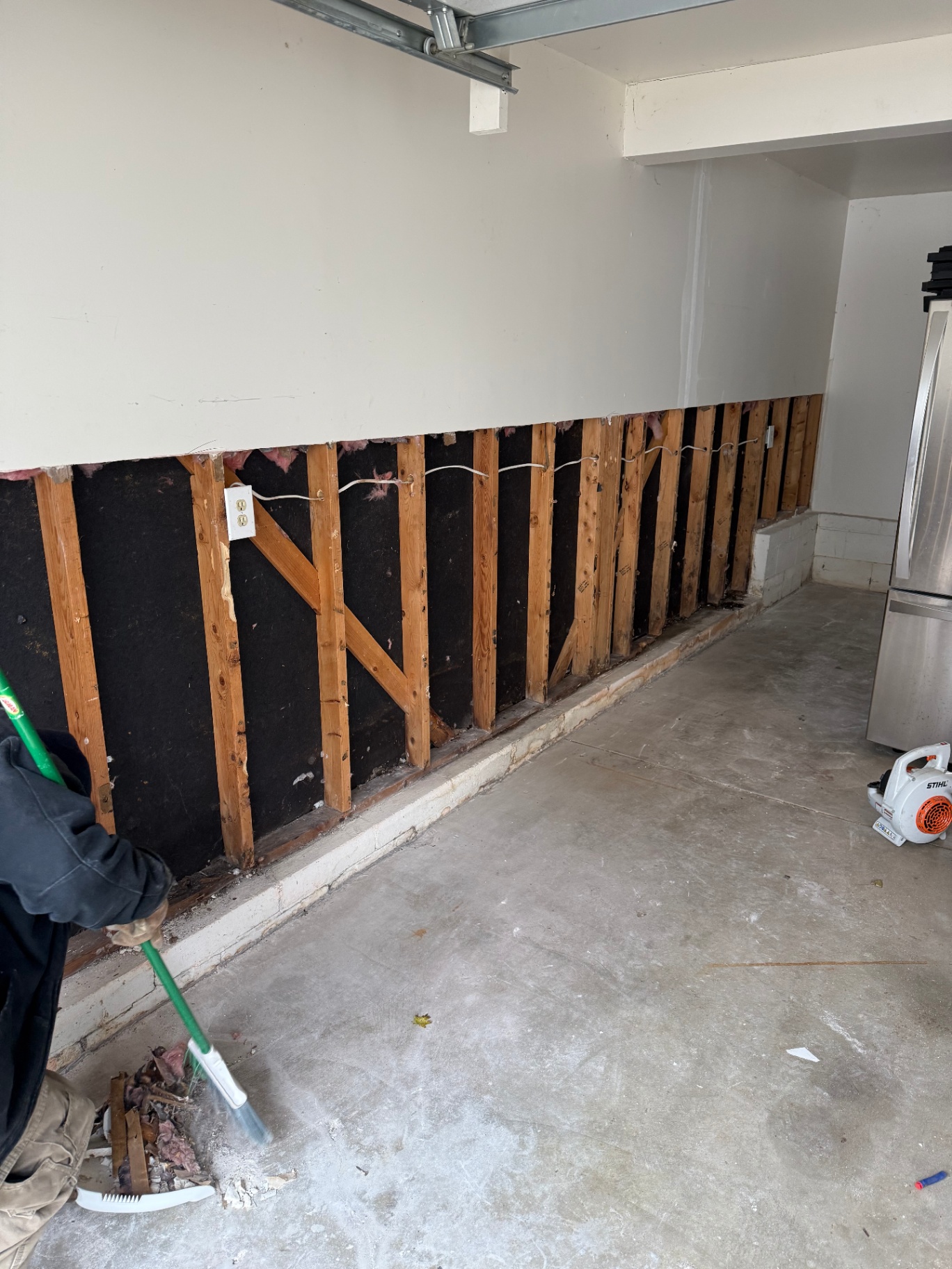 Long exposed run of garage wall with the lower drywall cut out to inspect framing, wiring, and the full extent of water damage